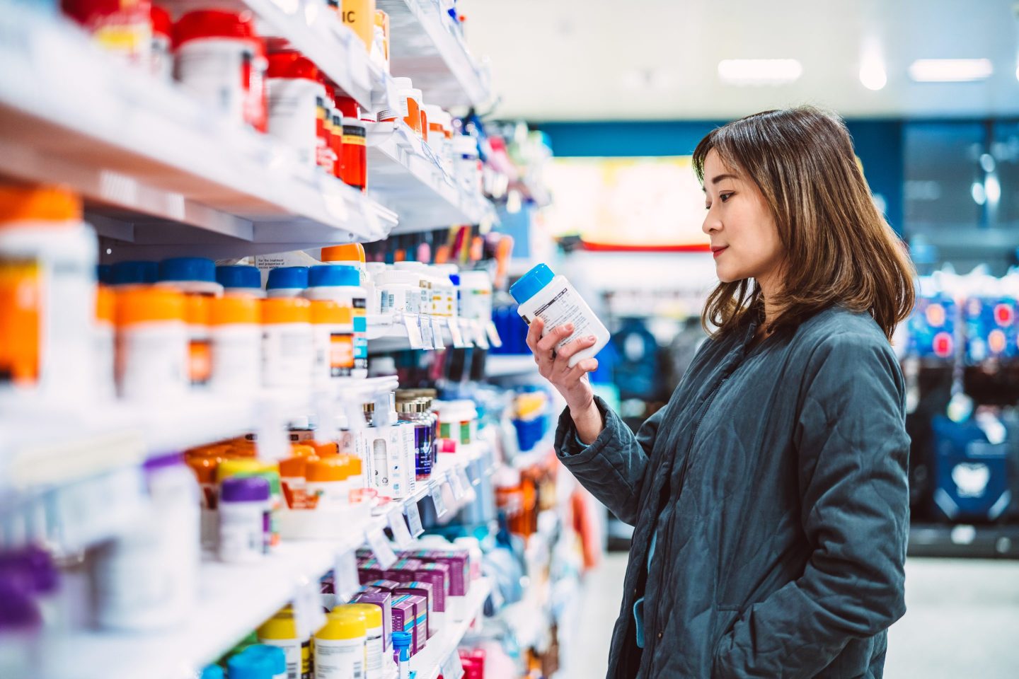 Beautiful Asian woman reading the ingredient label while shopping for multi vitamin &amp; health supplements on shelf in supermarket. Heathy lifestyle concept.